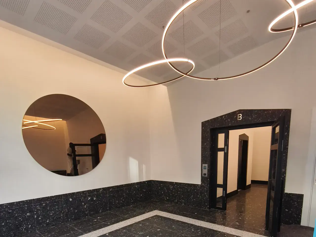 Hall de résidence livré avec sol en terrazzo sombre, miroir rond et luminaires annulaires