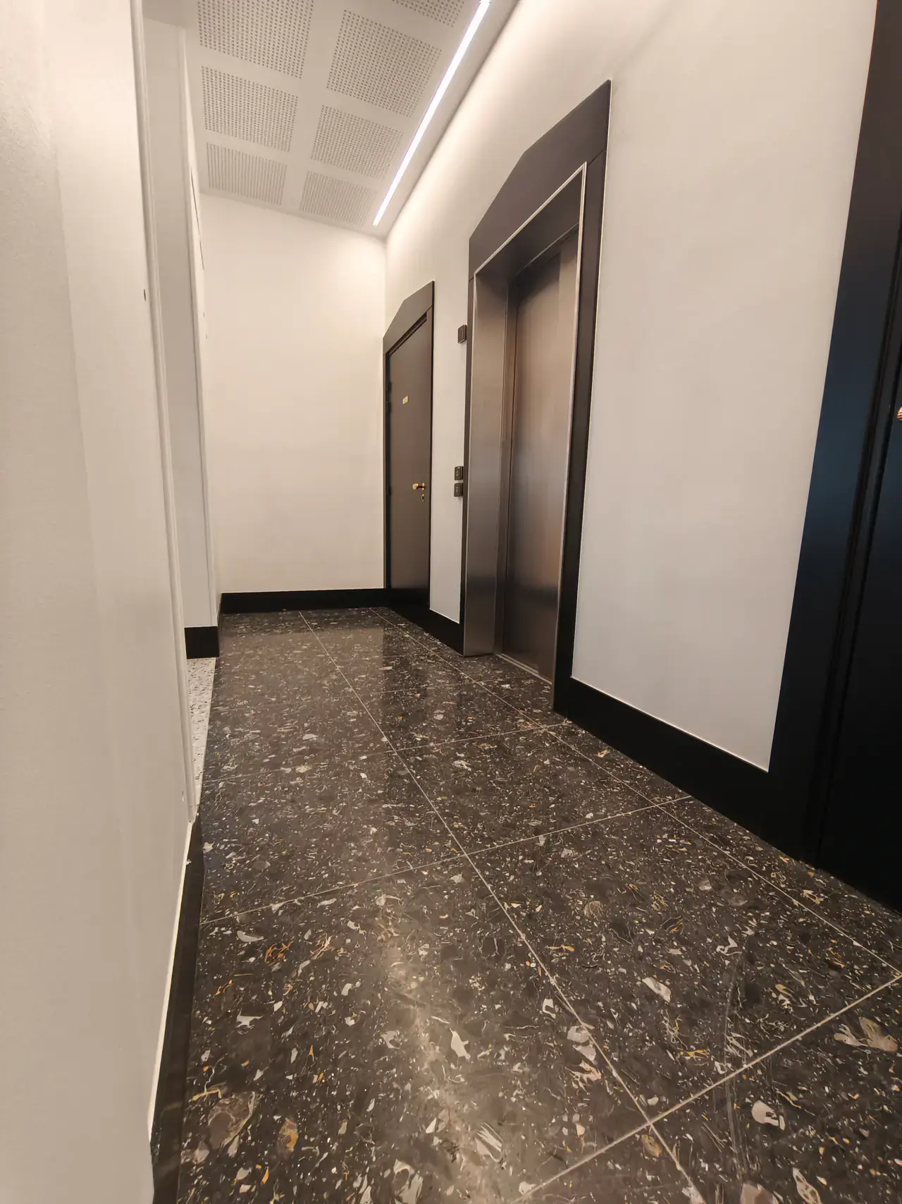 Palier de résidence livré avec sol en terrazzo sombre, murs clairs et porte d’ascenseur inox