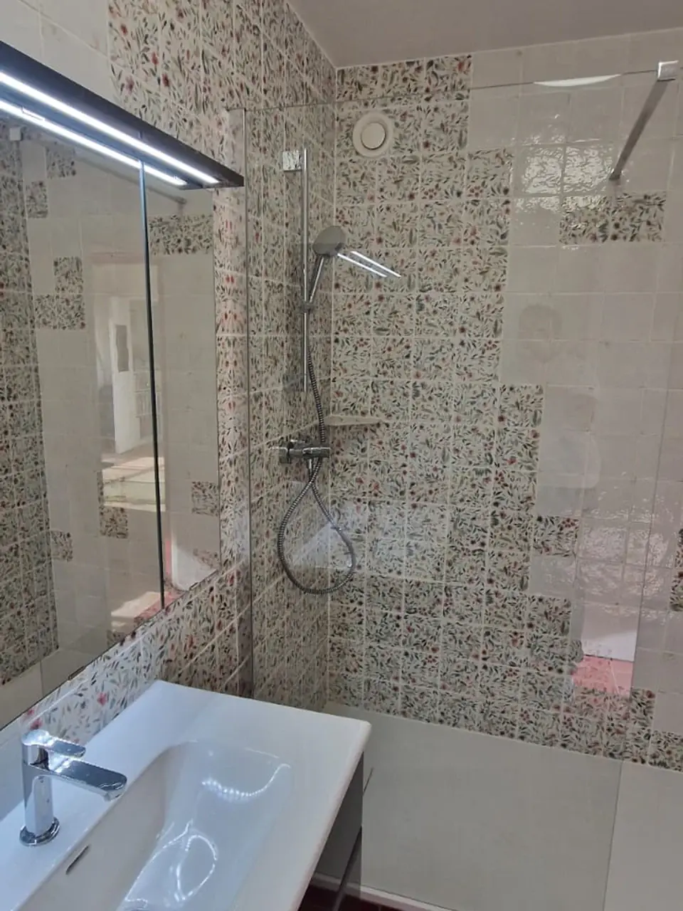 Meuble vasque sombre devant une douche vitrée et une faïence blanche à motifs fleuris