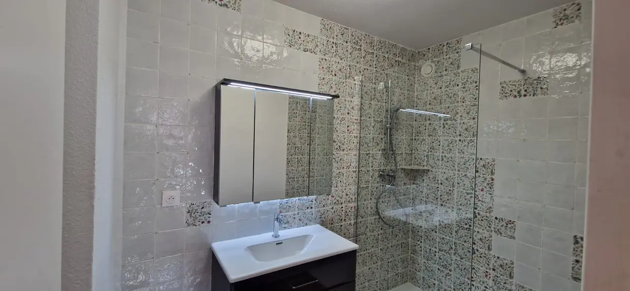 Salle de bains avec grande douche vitrée, meuble vasque et faïence blanche à motifs fleuris