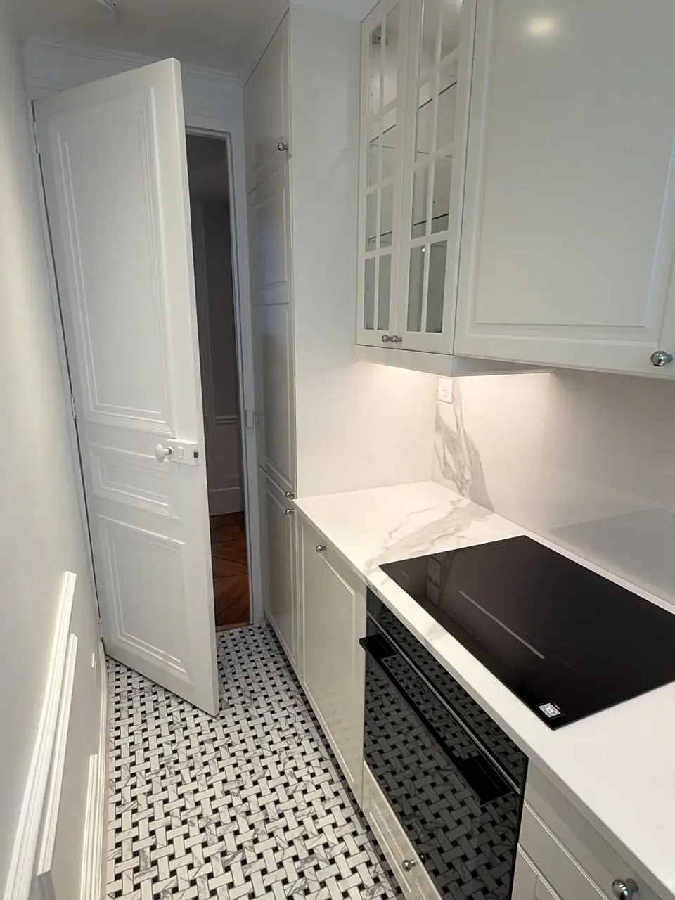 Cuisine étroite entièrement équipée avec meubles moulurés clairs, plan de travail effet marbre et sol en mosaïque